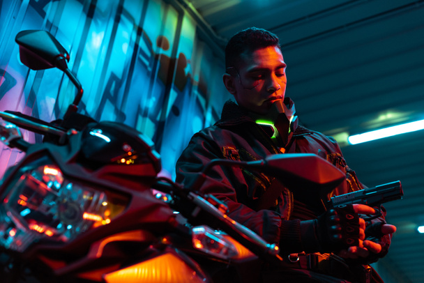 selektywny nacisk na bi-rasowy cyberpunk gracz w pobliżu motocykla patrząc na broń - Zdjęcie, obraz