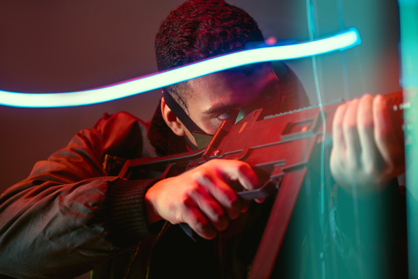 selective focus of bi-racial cyberpunk player in protective mask aiming gun near neon lighting on black  - Fotografie, Obrázek