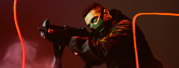 panoramic shot of bi-racial cyberpunk player in protective mask aiming gun near neon lighting on black  - Photo, Image