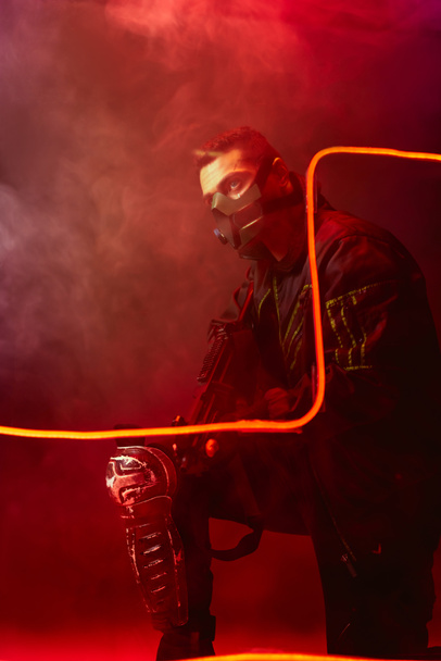 dangerous bi-racial cyberpunk player in protective mask holding gun near neon lighting on black with smoke  - Photo, Image