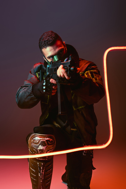 bi-racial cyberpunk man in mask aiming gun near neon lighting on black  - Zdjęcie, obraz