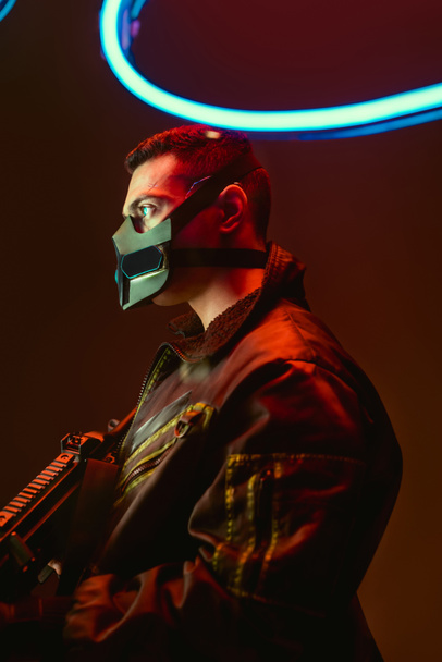 bi-racial cyberpunk player in mask holding gun near blue neon lighting on black  - 写真・画像