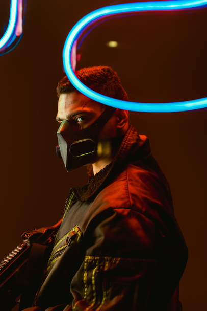selective focus of dangerous bi-racial cyberpunk player in mask holding gun near blue neon lighting on black  - Photo, Image