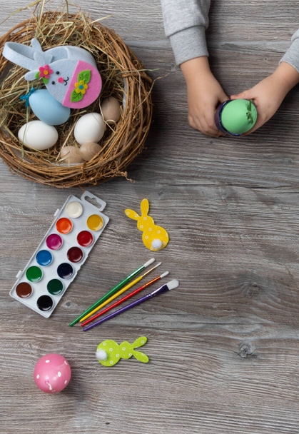 baby hand turn easter egg. In wooden table paints, brushes, bunny, nest. Prepares to paint Easte eggs - Zdjęcie, obraz