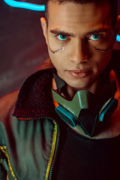 handsome and bi-racial cyberpunk player looking at camera near neon lighting on black  - 写真・画像