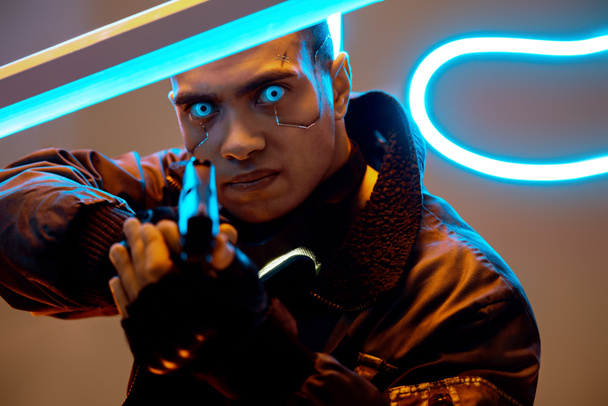 selektiver Fokus eines gutaussehenden und gemischten Cyber-Punk-Spielers mit metallischen Platten im Gesicht und blauen Augen, der eine Waffe in der Nähe von Neonlicht hält  - Foto, Bild