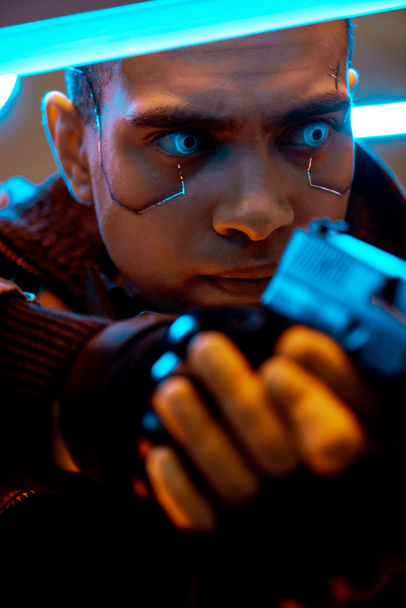 Selektiver Fokus eines birassischen Cyberpunk-Spielers mit metallischen Platten im Gesicht und blauen Augen, der eine Waffe in der Nähe von Neonlicht hält  - Foto, Bild