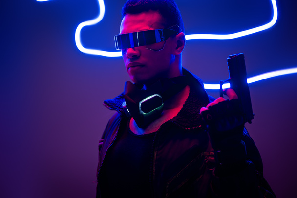μικτή φυλή Cyberpunk player σε φουτουριστικό γυαλιά κρατώντας όπλο κοντά σε μπλε νέον φωτισμό  - Φωτογραφία, εικόνα