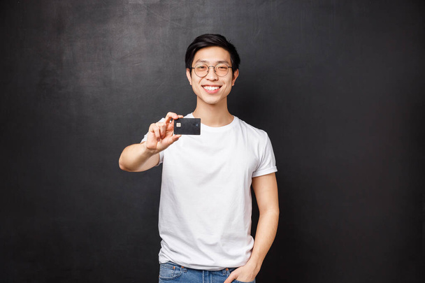 Bankovní, finanční a platební koncepce. Portrét přátelského sebevědomého mladého asijského muže ukazující kreditní kartu před kamerou s potěšeným zářivým úsměvem, rada otevřený účet na dát peníze, černé pozadí - Fotografie, Obrázek