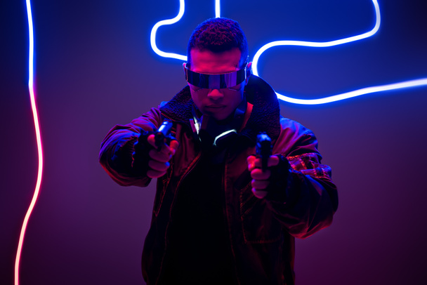 selective focus of mixed race cyberpunk player in futuristic glasses holding guns near neon lighting  - Фото, зображення