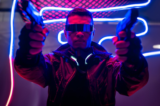 selective focus of armed mixed race cyberpunk player in futuristic glasses holding guns near neon lighting  - Photo, Image