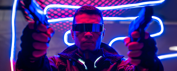 panoramic shot of mixed race cyberpunk player in futuristic glasses holding guns near neon lighting  - Foto, imagen