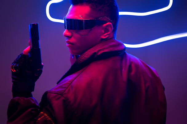 visão lateral do jogador cyberpunk de raça mista em óculos futuristas segurando arma perto de iluminação de néon
  - Foto, Imagem