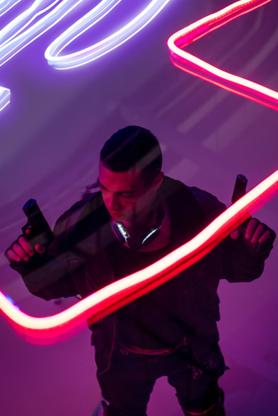 overhead view of armed bi-racial cyberpunk player holding guns near neon lighting  - Photo, Image