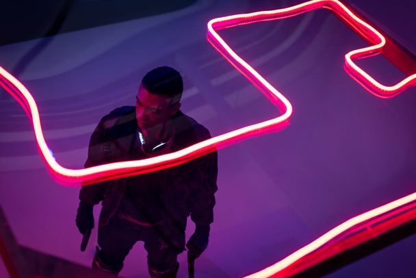 overhead view of armed bi-racial cyberpunk player holding guns near red neon lighting  - Photo, Image