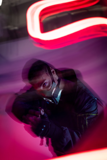 motion blur of armed bi-racial cyberpunk player in mask holding gun near red neon lighting  - Photo, Image