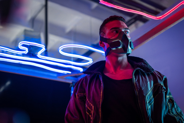 Mixed Race Cyberpunk-Spieler mit Maske steht in der Nähe von Neonlicht und schaut weg - Foto, Bild