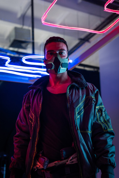 jugador cyberpunk de raza mixta en máscara de pie cerca de la iluminación de neón
  - Foto, imagen