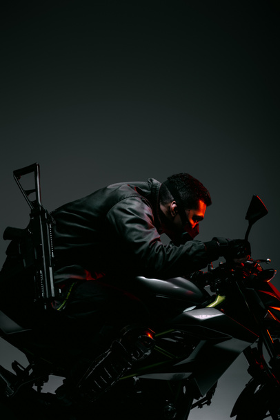 profile of armed mixed race cyberpunk player in mask riding motorcycle on grey  - Photo, Image