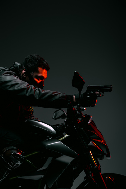Seitenansicht des bewaffneten bi-rassischen Cyberpunk-Spielers in Maske, der Motorrad fährt und Pistole auf grau hält  - Foto, Bild