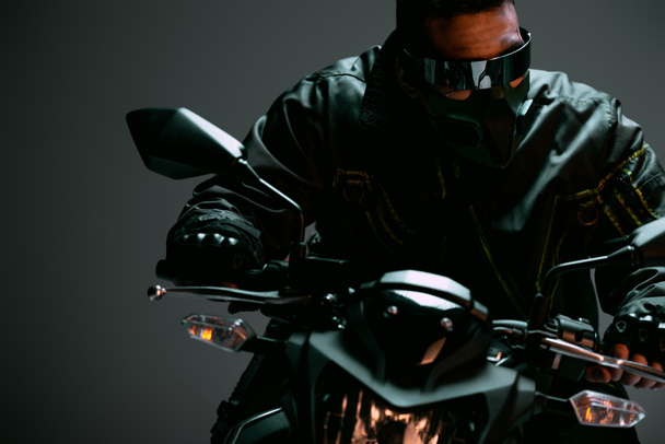 selective focus of bi-racial cyberpunk man in mask and futuristic glasses riding motorcycle on grey  - Фото, изображение