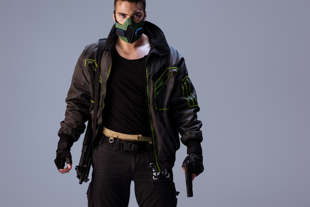 ένοπλη μικτή φυλή cyberpunk player σε μάσκα στέκεται απομονωμένο σε γκρι   - Φωτογραφία, εικόνα