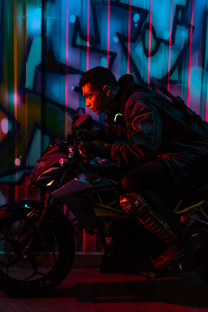side view of mixed race cyberpunk player riding motorcycle on street with graffiti - Foto, afbeelding