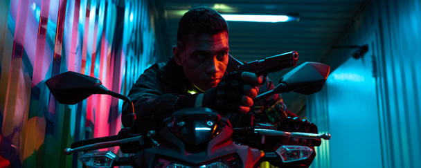 panoramisch schot van bi-raciale cyberpunk speler op motorfiets gericht pistool op straat met graffiti - Foto, afbeelding