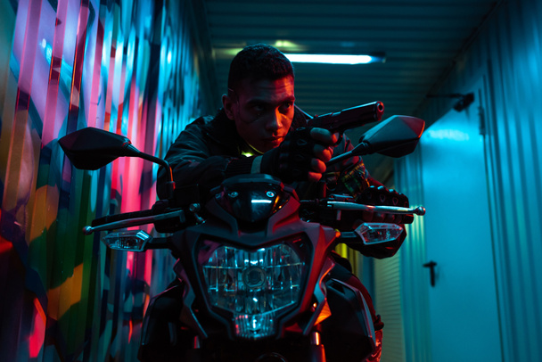 bi-rasowy cyberpunk gracz na motocyklu celuje pistolet na ulicy z graffiti - Zdjęcie, obraz