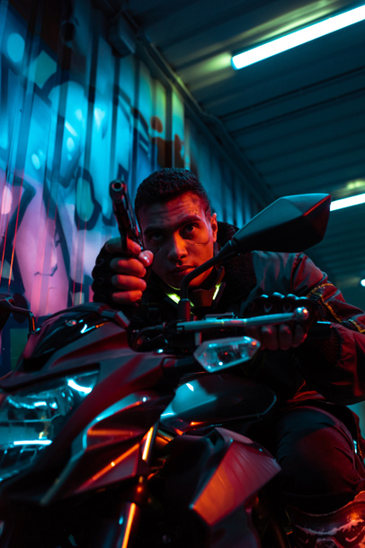 selective focus of mixed race cyberpunk player on motorcycle aiming gun on street with graffiti - Photo, Image