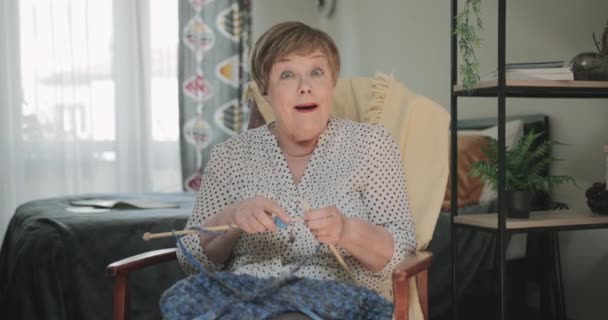 Old cheerful lady sitting on chair and knitting at her apartment. Front view of elderly woman looking surprised while holding knitting needles and smiling to camera.Concept of leisure. - Metraje, vídeo