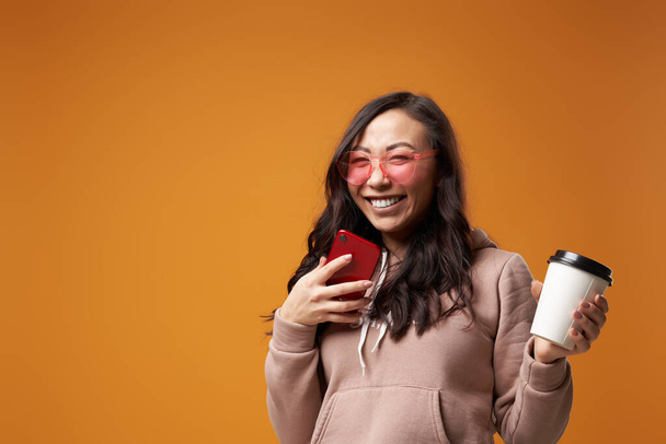 Happy asian woman in pink glasses with glass and phone in her hands in studio - Photo, image