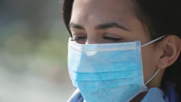 Young brunette woman standing in a blue protective mask outdoors in spring         Amazing closeup of a stylish brunette girl with long loose hair waving in windy weather standing in a protective mask against coronavirus on the Dnipro bank in spring - Materiaali, video
