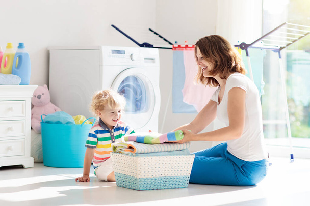 Mutter und Kinder in Waschküche mit Waschmaschine oder Wäschetrockner. Hausarbeit. moderne Haushaltsgeräte und Waschmittel in weißen, sonnigen Wohnungen. sauber gewaschene Kleidung auf dem Wäscheständer.  - Foto, Bild