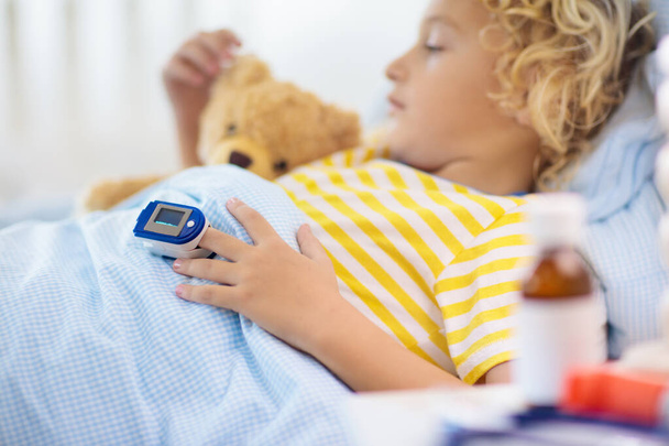 Sick little boy with pulse oximeter on his finger. Asthma treatment. Ill child lying in bed. Unwell kid with chamber inhaler, cough medicine. Flu season. Bedroom or hospital room for young patient.  - Photo, Image