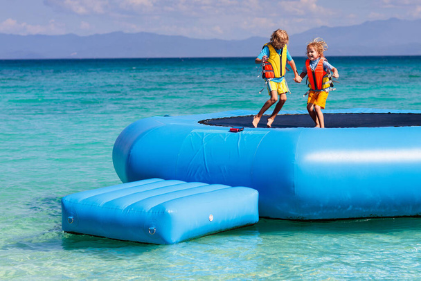 熱帯の海のビーチでトランポリンでジャンプする子供たち。子供たちは膨脹可能なウォータースライドでジャンプします。エキゾチックな島のリゾートでアクアアミューズメントパーク。家族での休暇と子供との旅行。海沿岸の楽しみ. - 写真・画像
