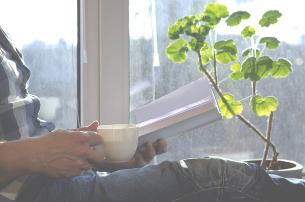 Ένας άνθρωπος κάθεται σε ένα περβάζι παραθύρου με ένα φλιτζάνι καφέ και ένα βιβλίο κατά τη διάρκεια της καραντίνας λόγω της πανδημίας του coronavirus - Φωτογραφία, εικόνα