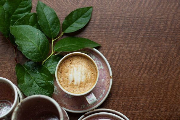 A cup of coffee with milk foam. Cappuccino stands among other dishes of the same color on the table. A branch of greenery decorates the composition. Homeliness and pleasant atmosphere. Top View. - Foto, Bild