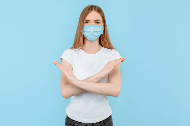A young woman, wearing a protective medical mask , shows a stop gesture with crossed arms, air pollution. Protection against the Chinese coronavirus pandemic, on an isolated blue background - Foto, Imagen