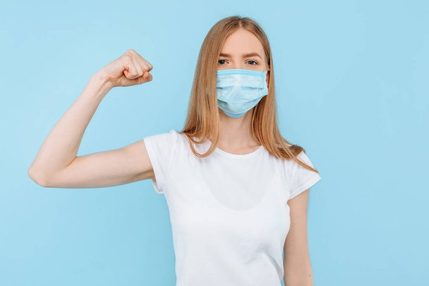 Una joven con una máscara médica protectora en la cara muestra un gesto de fuerza, sobre un fondo azul aislado. Contaminación atmosférica, virus, coronavirus
 - Foto, Imagen