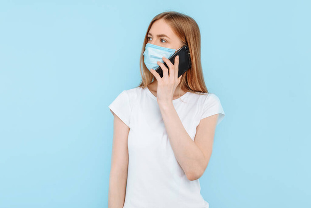 Uma jovem mulher usando uma máscara médica protetora em seu rosto está falando em um telefone celular, de pé contra um fundo azul isolado. Poluição do ar, vírus, coronavírus
 - Foto, Imagem