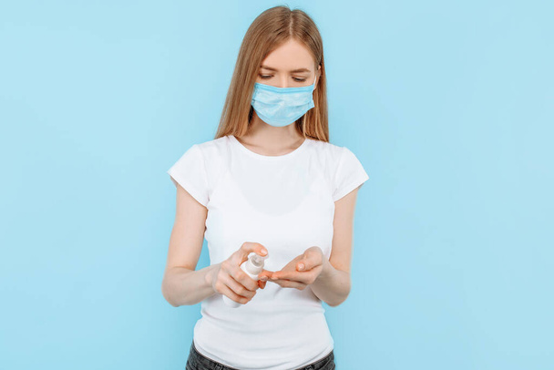 A young girl in a protective medical mask on her face, holding a disinfectant, hand antiseptic, on an isolated blue background. Concept of a virus, coronavirus - Фото, зображення