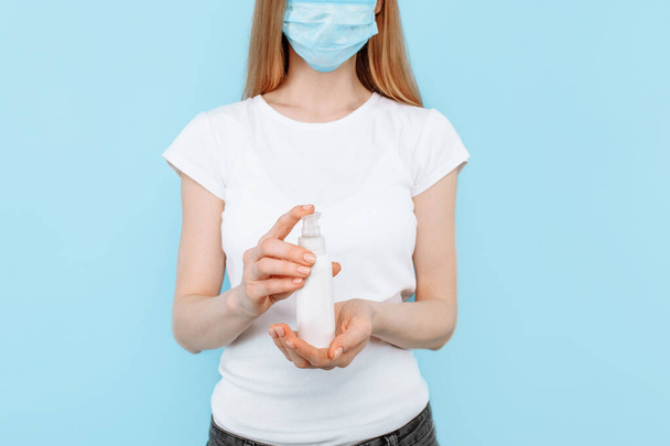 Een jong meisje met een beschermend medisch masker op haar gezicht, met een ontsmettingsmiddel, met de hand ontsmettend, op een geïsoleerde blauwe achtergrond. Concept van een virus, coronavirus - Foto, afbeelding