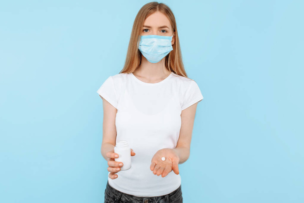 Una joven con una máscara médica protectora en la cara, sosteniendo un frasco de pastillas, sobre un fondo azul aislado. Concepto de virus o coronavirus
 - Foto, imagen