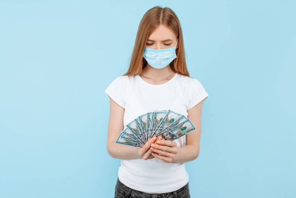 girl in a protective medical mask on her face, holding cash dollar bills, on an isolated blue background. concept of a virus , a coronavirus , rising prices for medical masks, expensive medical care - Photo, image