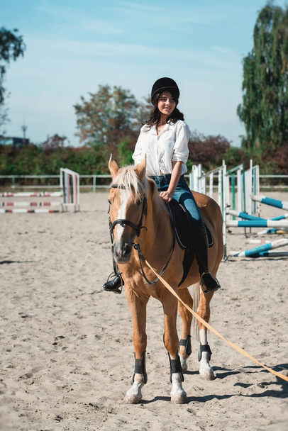 Muotokuva nuori brunette nainen valkoinen paita ja kypärä hevosen selässä. oppii ratsastamaan. ratsastus oppitunteja hevonen epodrom
 - Valokuva, kuva