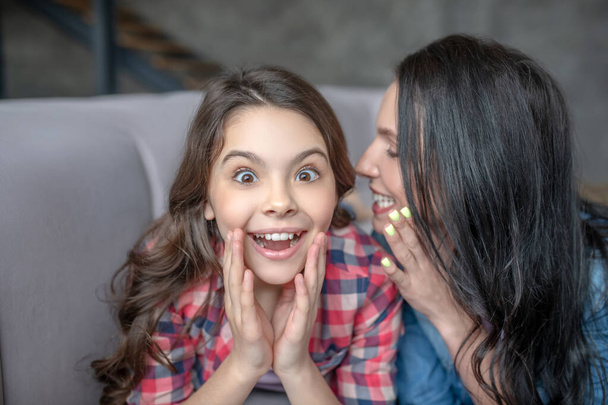 Dark-haired mom telling something interesting to her surprised daughter in the ear - Foto, imagen