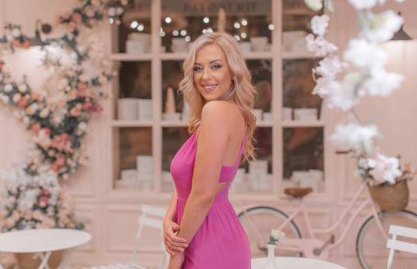 Atraktivní šťastná blondýna v růžové sexy šaty pózovat mezi místy stylizované jako pařížská kavárna zdobené kudrnaté květiny a vinobraní kolo - Fotografie, Obrázek
