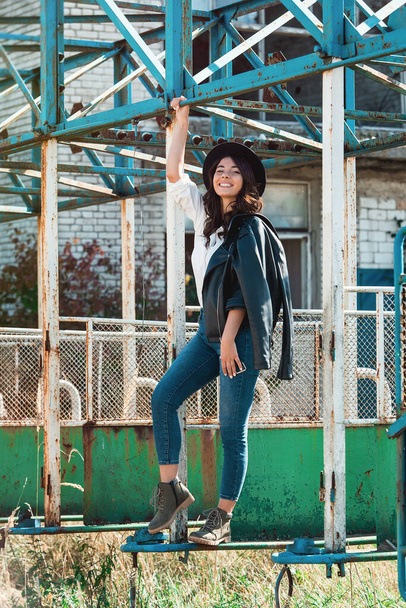 Beautiful sexy girl posing on the rustic metal construction. Female wearing white shirt, black hat and blue denim jeans. black jacket on the shoulder - Фото, зображення