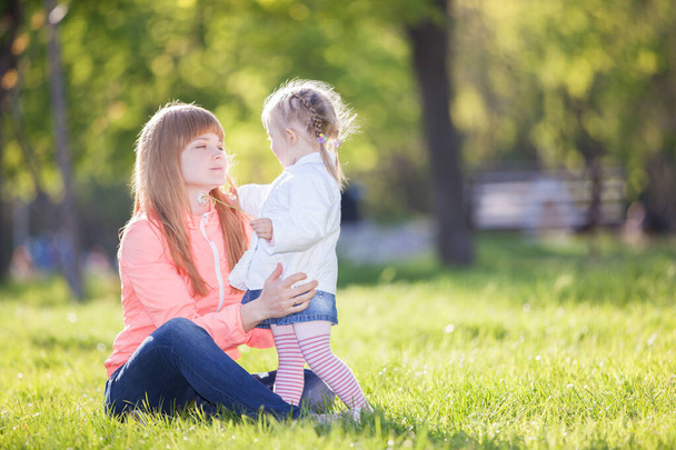 Mother and daughter in the park. Beauty nature scene with colorful background at spring season. Family outdoor lifestyle. Happy woman and cute girl relax on green grass - Zdjęcie, obraz